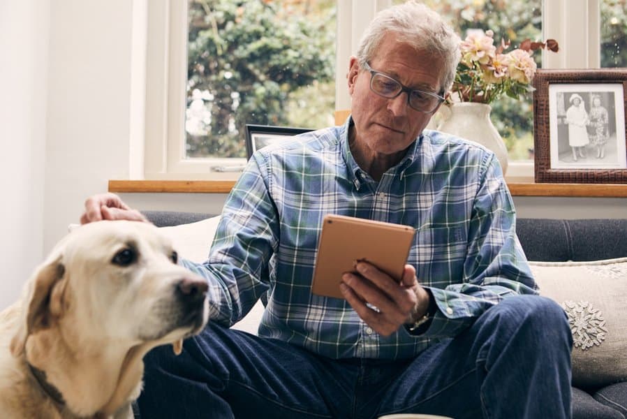 An older man looking at a tablet in his living room, with his dog by his side