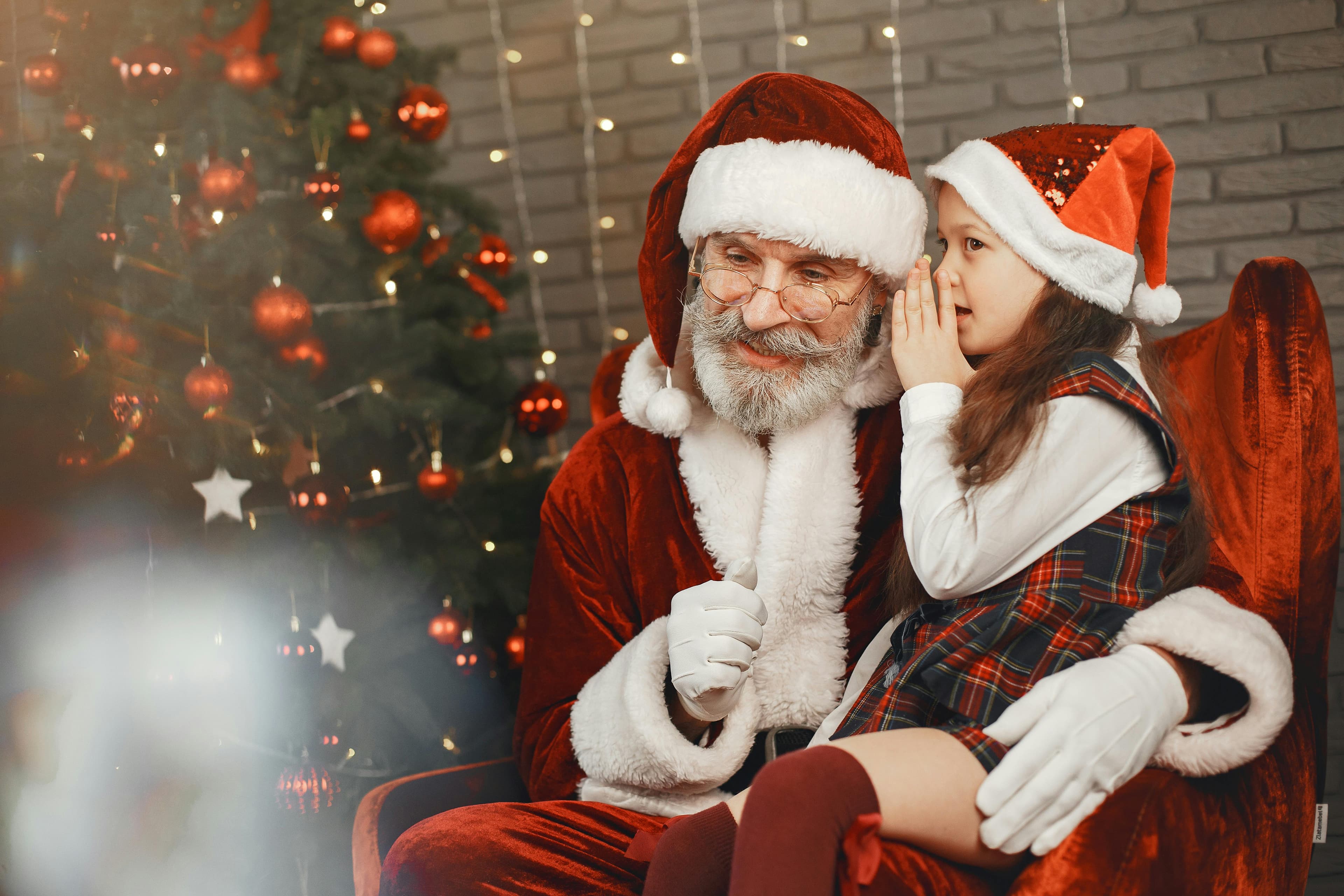 Free Santa Claus sharing a secret with a child by the Christmas tree, surrounded by festive decorations. Stock Photo
